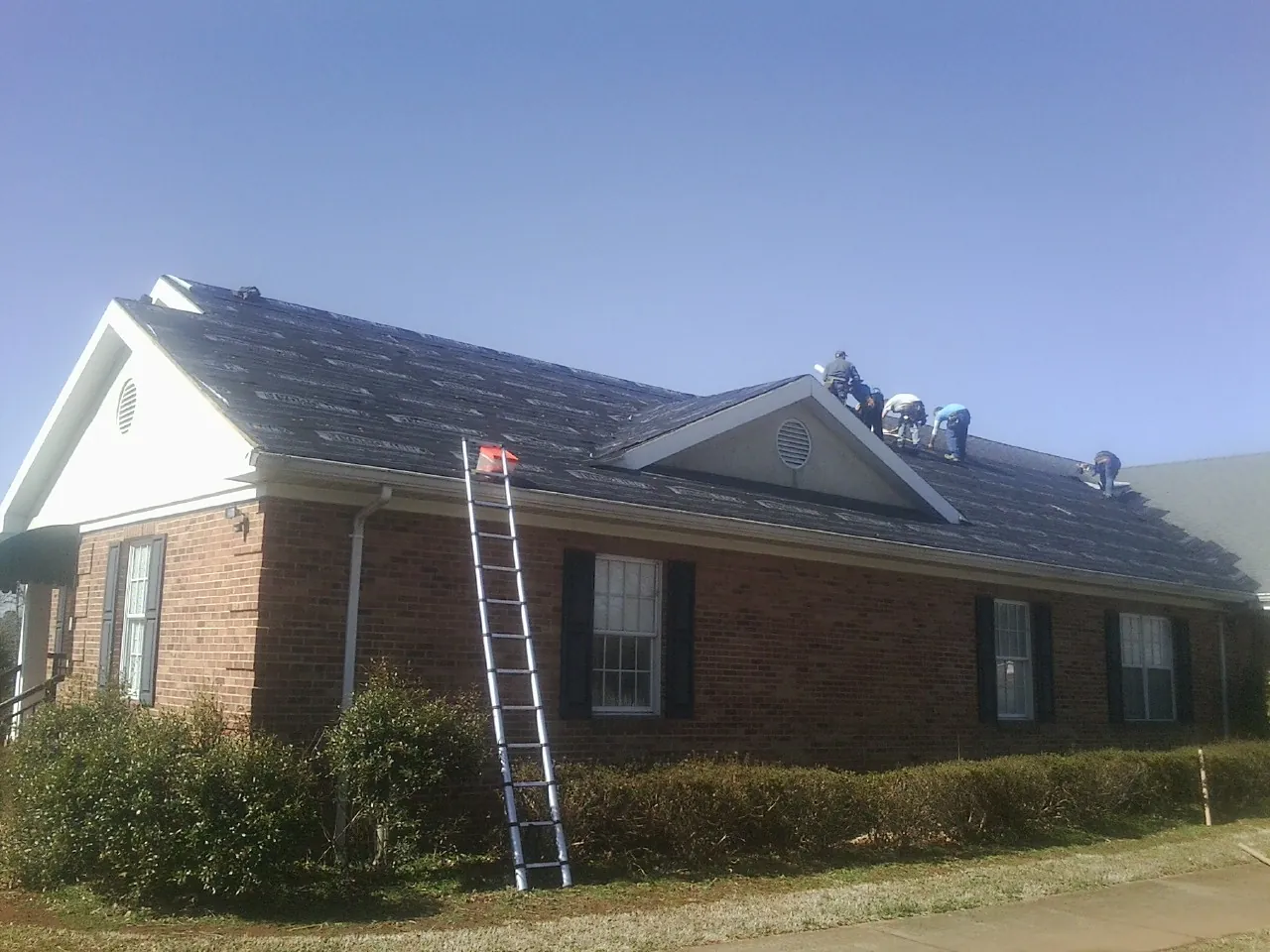 Crew tearing shingles off a brick ranch roof with a ladder set — Emergency Roof Repair work by our crew in Victor